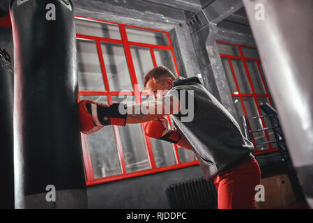 Fiducioso giovane sportivo muscolare in rosso guanti per sport esercita sul punzone pesante in borsa. Duro allenamento in palestra di inscatolamento sfondo Foto Stock