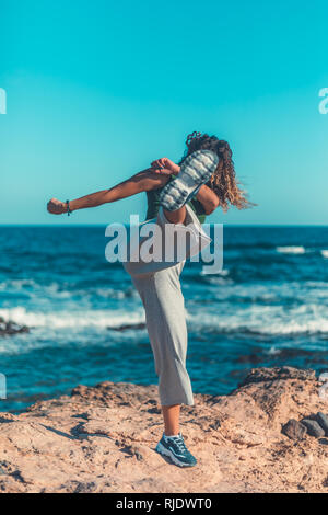 Irriconoscibile femmina in abito casual calci aria mentre in piedi sulla riva sassosa contro agitando il mare e cielo blu senza nuvole Foto Stock