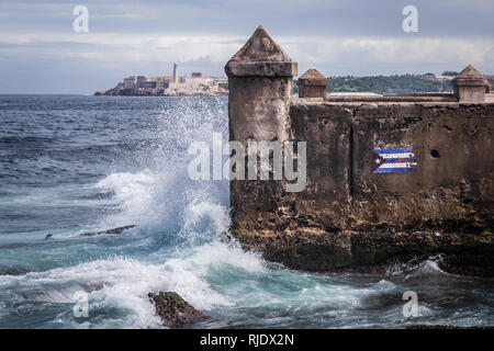 Le onde battono su un interruttore d'onda con un disegno della bandiera cubana sul Malecon promenade a l'Avana, Cuba. Morro castle e un faro sono visibili dietro Foto Stock