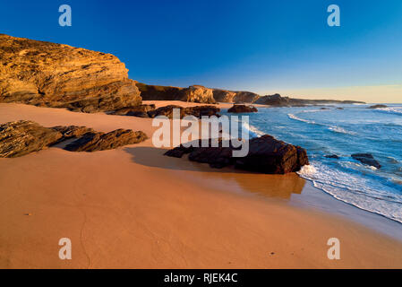 Lonely spiaggia con sabbia e rocce fodera una calma blu oceano con onde morbide Foto Stock