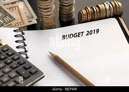 Pianificazione del bilancio concetto. Blocco note con il bilancio 2019 testo, matita, calcolatrice e dollaro banconote, pile di monete sullo sfondo Foto Stock