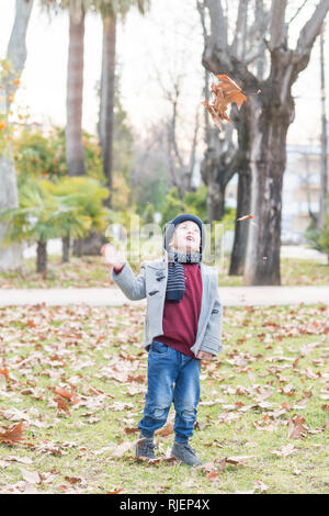 Bambino vestito in abiti caldi gettando caduto foglie di autunno in aria Foto Stock