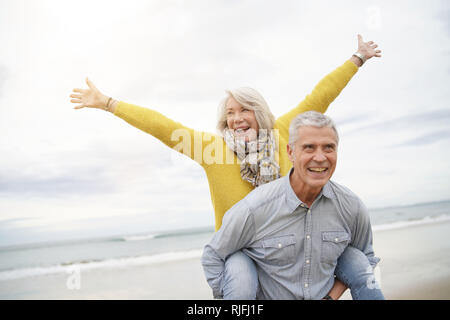 Moderno e vivace coppia senior piggy back riding sulla spiaggia Foto Stock