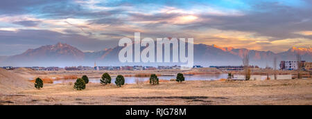 Il lago e le case contro la montagna soleggiati in Alba Foto Stock