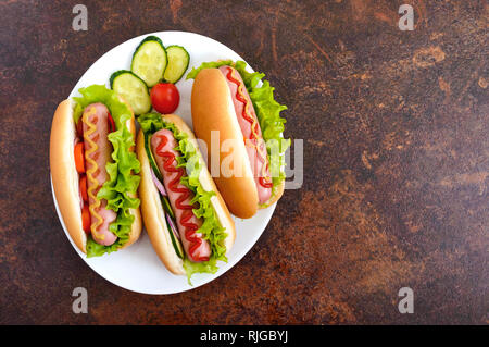 Gustosi piatti caldi i cani con la salsiccia grill, ketchup, senape, lattuga, pomodori su una piastra bianca. La vista dall'alto. Lay piatto Foto Stock