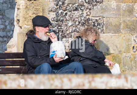 Coppia senior seduti fuori in inverno nel Regno Unito a mangiare pesce e patatine da un sacchetto di carta. Foto Stock