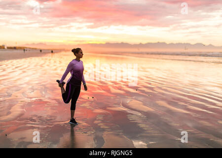 Giovani femmine pareggiatore di stretching prima di correre sulla spiaggia di sunrise Foto Stock