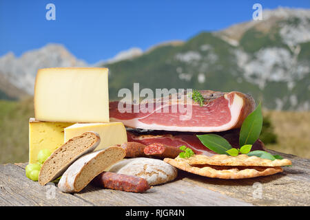 Specialità sudtirolesi come bacon, salsicce e formaggi giacenti su un tavolo rustico di fronte montagne delle Alpi Foto Stock