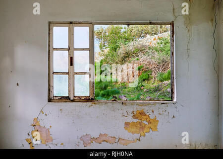 Interno di una casa in rovina con vecchio e sporco e screpolata muro bianco e la rottura di un telaio di finestra che si affaccia sul prato di view Foto Stock