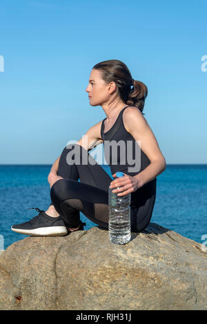 Sportive seduto su una roccia con una bottiglia di plastica di acqua in appoggio sul mare dopo il corso di formazione e di esercizio. Foto Stock