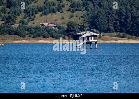 Haunted abbandonato ex casa di partito nelle calme acque blu del serbatoio acqua Dospat nella Bulgaria meridionale. Tetto bianco di escrementi di uccelli. Diversi i cormorani in appoggio sulla parte superiore. Soleggiata giornata autunnale Foto Stock