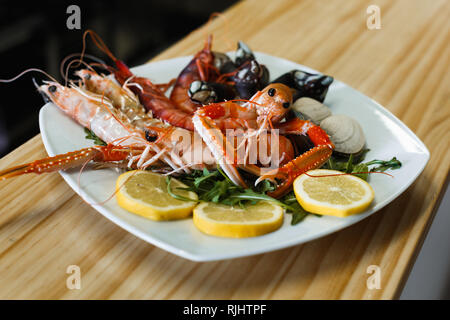 Piastra di colore bianco con gamberi cotti sgusciati, gli scampi e le vongole con le erbe e le fette di limone su sfondo di legno Foto Stock