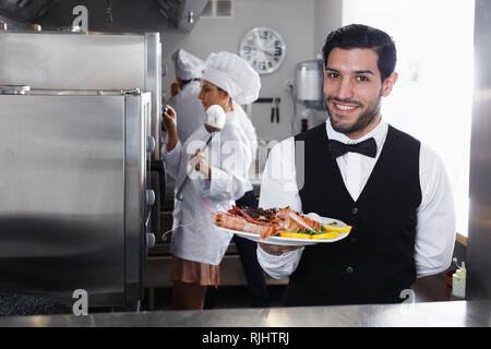 Ritratto di bello cameriere con piatto di frutti di mare in cucina in un ristorante di pesce Foto Stock