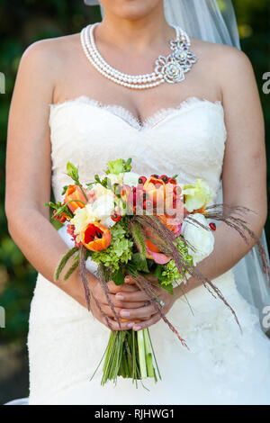 Sposa tenendo il suo matrimonio bouquet di fiori con verde, rosso, bianco e fiori d'arancio Foto Stock