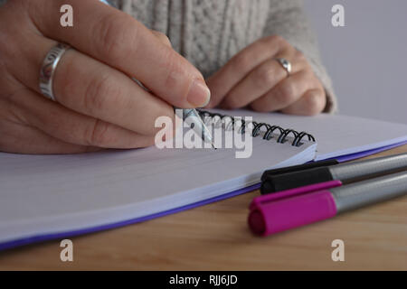 Donna iscritto in una rilegatura a spirale notebook, penna in mano, in bilico su una pagina vuota Foto Stock