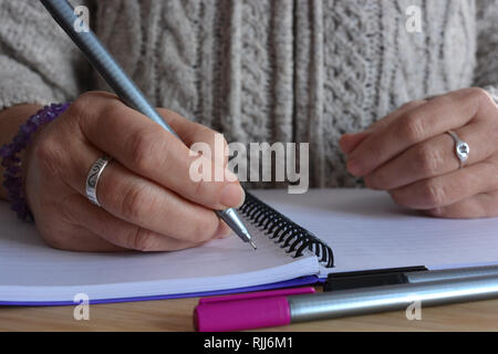 Donna iscritto in una rilegatura a spirale notebook, penna in mano, in bilico su una pagina vuota Foto Stock