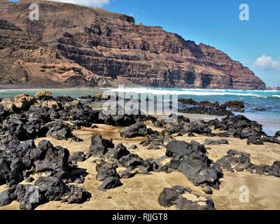 La Lava sulla spiaggia sabbiosa a Orzola, Lanzarote nelle Isole Canarie con scogliere di sfondo e il blu delle onde dell'Atlantico Foto Stock