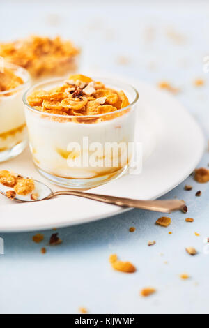 Sana colazione o dessert. Gustosi corn flakes con yogurt, mandorla e miele in un bicchiere piccolo e un cucchiaio di yogurt e fiocchi di mais su bianco che serve Foto Stock