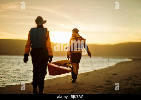 Felice coppia senior che trasportano un kayak insieme lungo una spiaggia di sabbia. Foto Stock