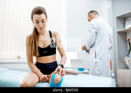 Giovane donna seduta del paziente con kinesio nastro sul suo ginocchio durante il trattamento medicale con terapista in ufficio Foto Stock