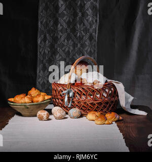 Cesto di pasqua con il cibo e la ciotola con ciambelle, gli uccelli e le uova sono sul tavolo da bassa illuminazione chiave contro il rito ucraino panno. Foto Stock