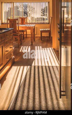 La luce del sole splende in una cucina attraverso veneziane verticali la colata di una lunga ombra a strisce sul pavimento in legno e di tutta la quercia tavolo da cucina e sedie Foto Stock