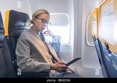Bionda caucasica casual donna che indossa gli occhiali lettura digitale sul lettore di posta elettronica mentre viaggiano in aereo. Trasporto commerciale da piani. Foto Stock