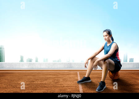 Felice donna asiatica con sport jersey seduta sul basket all'aperto in campo di pallacanestro Foto Stock