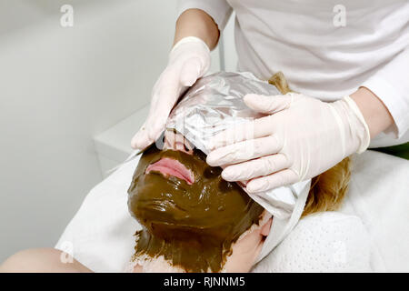 Estetista lavoratore mette un foglio di alluminio sul cliente femmina faccia, procedura dopo l'applicazione di cosmetici per il viso maschera di mare ​​algae per anti invecchiamento Foto Stock