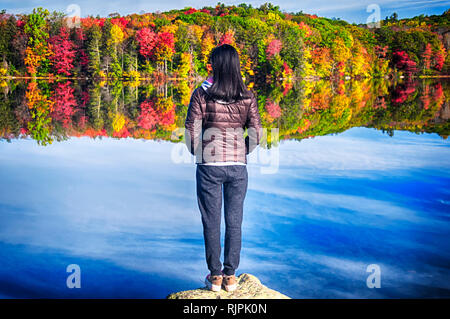 Una donna cinese in piedi su una roccia a guardare la caduta delle foglie che riflette in acqua mentre è seduto su un tavolo da picnic in autunno a Burr Pond State pa Foto Stock