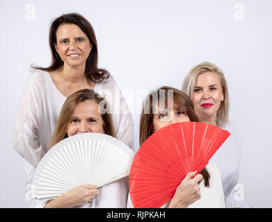 Gruppo di 4 donne, amici di mezza età divertendosi in una sessione di foto in uno studio con sfondo bianco, seduto su una palla e giocando con una ventola Foto Stock