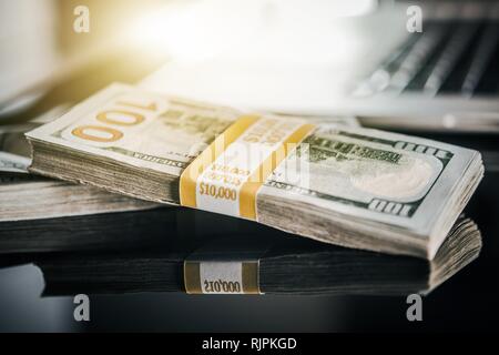 Tema di business. Denaro contante stipendio. Dieci di migliaia di dollari di pile sulla scrivania vetroso con computer portatile in background. Foto Stock
