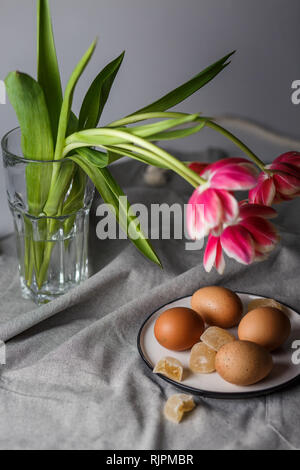 Bollite le uova di colore marrone su una piastra vicino ai tulipani in un vetro trasparente cup su sfondo grigio in una luce ovattato e in soft focus Foto Stock