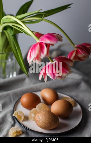 Bollite le uova di colore marrone su una piastra vicino ai tulipani in un vetro trasparente cup su sfondo grigio in una luce ovattato e in soft focus Foto Stock