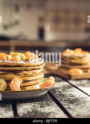 Foto verticale - pila di frittelle dorate con banane e arance sulla tavola di legno ricoperta di zucchero semolato. Cumulo di frittelle americane Foto Stock