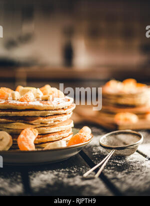 Foto verticale - pila di frittelle dorate con banane e arance sulla tavola di legno ricoperta di zucchero semolato. Cumulo di frittelle americane Foto Stock