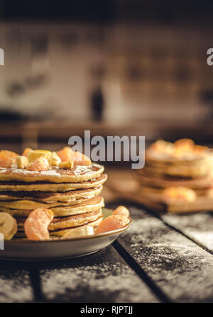 Foto verticale - pila di frittelle dorate con banane e arance sulla tavola di legno ricoperta di zucchero semolato. Cumulo di frittelle americane Foto Stock