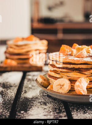 Foto verticale - pila di frittelle dorate con banane e arance sulla tavola di legno ricoperta di zucchero semolato. Cumulo di frittelle americane Foto Stock