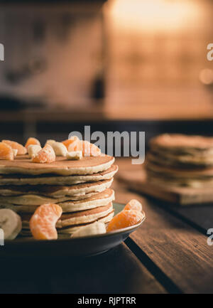 Foto verticale - pila di frittelle dorate con banane e arance sulla tavola di legno ricoperta di zucchero semolato. Cumulo di frittelle americane Foto Stock