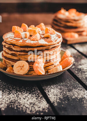 Foto verticale - pila di frittelle dorate con banane e arance sulla tavola di legno ricoperta di zucchero semolato. Cumulo di frittelle americane. Foto Stock