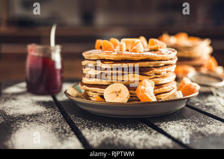 Pila di frittelle dorate con banane e arance sulla tavola di legno ricoperta di zucchero semolato. Cumulo di american pancake con sciroppo d'acero Foto Stock