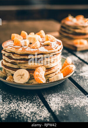 Foto verticale - pila di frittelle dorate con banane e arance sulla tavola di legno ricoperta di zucchero semolato. Cumulo di frittelle americane. Foto Stock