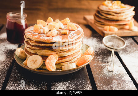 Pila di frittelle dorate con banane e arance sulla tavola di legno ricoperta di zucchero semolato. Cumulo di american pancake con sciroppo d'acero Foto Stock