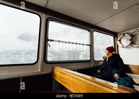 Woman in Red Hat seduti all'interno dei traghetti passeggeri sul modo di Hallstatt, Austria. In inverno la vista sul lago e le montagne delle Alpi. Foto Stock