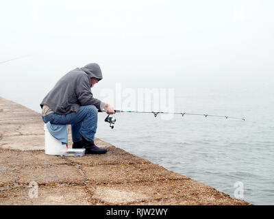 Un vecchio uomo in jeans blu e grigio con cappuccio Felpa si siede su una benna da 5 galloni mentre la pesca in un giorno di nebbia. Sud Jetty Port Aransas, Texas, Stati Uniti d'America. Foto Stock