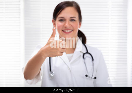 Vista frontale di Felice medico donna che mostra un pollice alzato in piedi in clinica Foto Stock