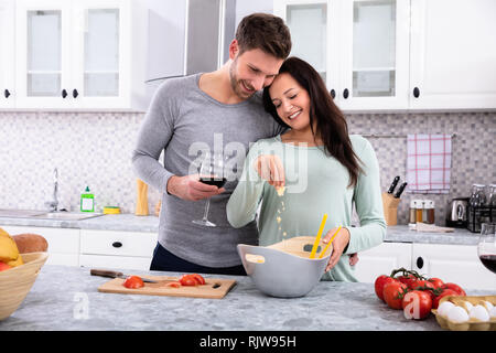 Sorridente moglie spargendo il burro con il marito in piedi in cucina Azienda bicchiere da vino Foto Stock