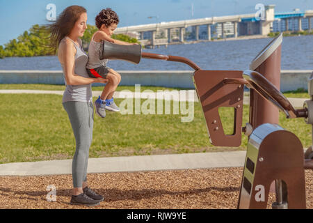 Una madre aiuta il suo bambino fino a usare la spalla per adulti tamponi di supporto sulla macchina di allenamento al Waterfront Park palestra. Foto Stock