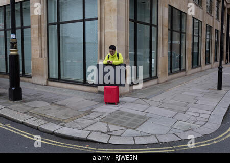 Un uomo in un verde lime e ponticello con red trolley bagaglio, utilizza il suo telefono cellulare su un angolo di strada nel centro di Londra, il 5 gennaio 2019, a Londra, in Inghilterra. Foto Stock