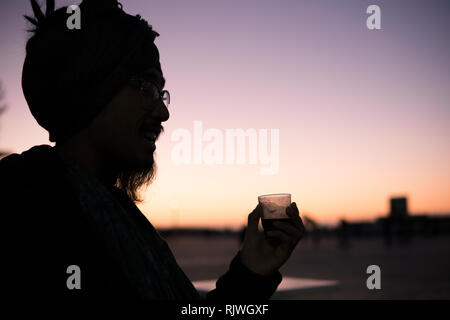 Giovani asiatici barbuto uomo turistica con Dreadlocks e sciarpa sulla testa Morroccan bere il caffè in Essaouira, Marocco al tramonto di sera tempo. Foto Stock